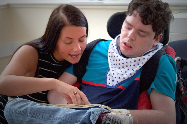 Next Step Trust a young woman points to a page in a book which sits on the lap of a young person in a wheelchair